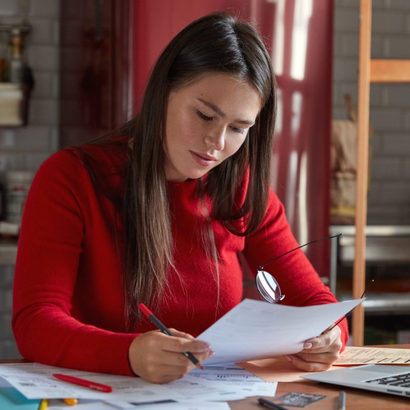 Woman chief architect works with documentation, confirms project, holds paper and glasses in hands, poses at kitchen, chats with marketing experts on laptop computer. People and career concept